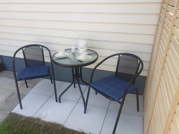 Une petite terrasse extérieure avec une table ronde et deux chaises, un pichet blanc et une théière blanche posés sur la table.
