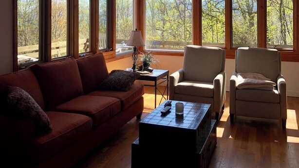 Una habitación amplia con ventanas grandes, un ventilador de techo, un sofá rojo, dos sillones y una mesa de centro.
