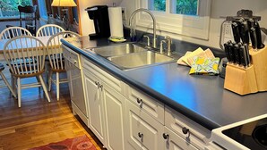 Una cocina con gabinetes blancos, un lavamanos doble y una estufa. Hay una mesa de comedor de madera con sillas, un tapete rojo y una ventana con vistas a la vegetación.