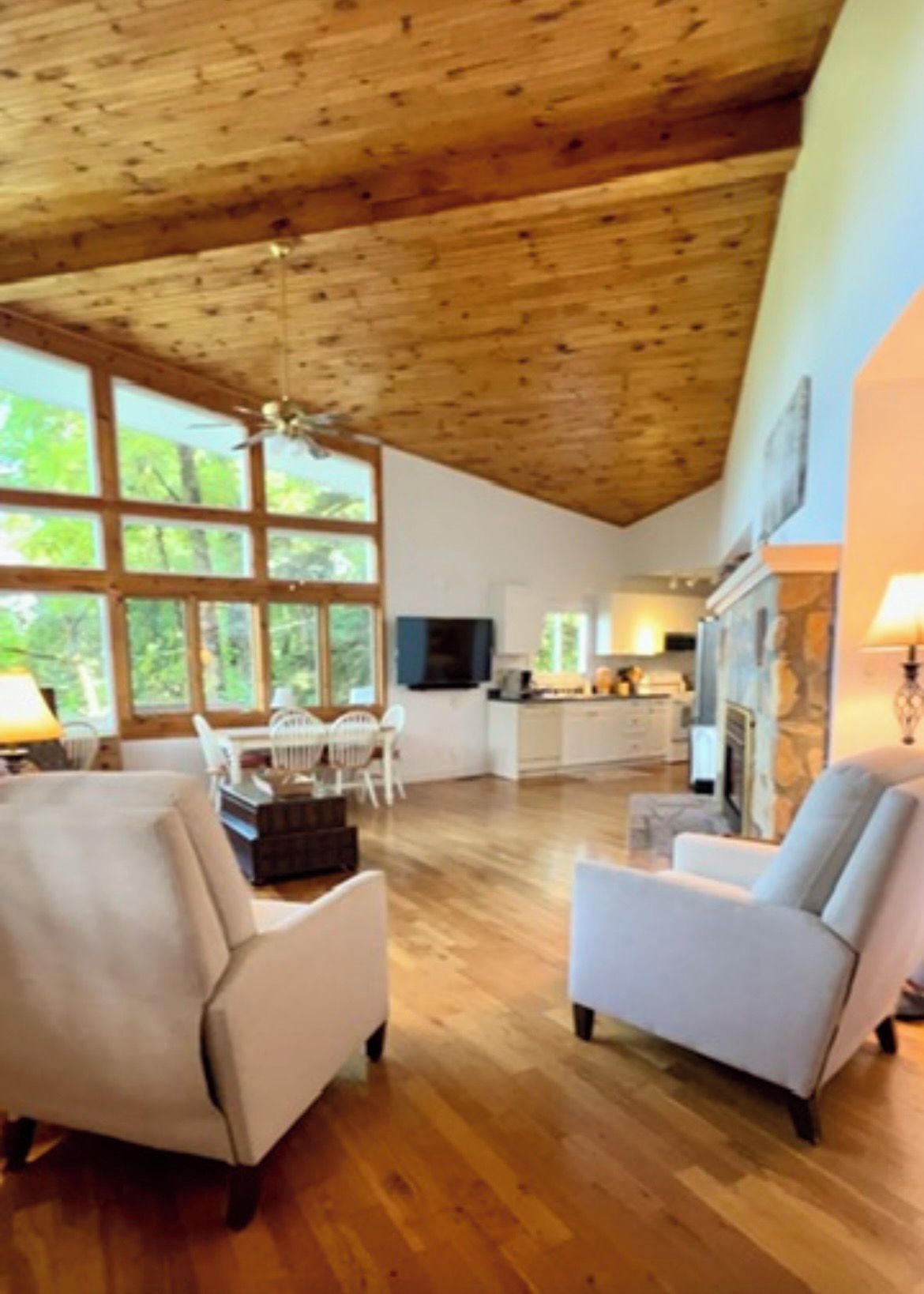 A spacious living room with a high ceiling and wooden beams, a fireplace, and a view of the outdoors through large windows.