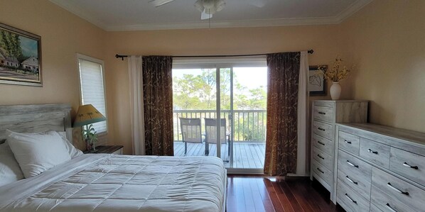 A bedroom with a bed, a dresser, a nightstand, a window with curtains, a ceiling fan, and a balcony with chairs.