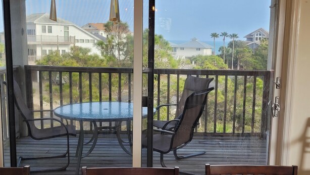 A balcony with a glass table and chairs, overlooking a residential area with houses and palm trees.