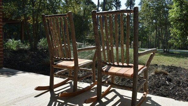 Two wooden rocking chairs on a concrete patio with a forest background. - YMCA Camp Cullen (Trinity)