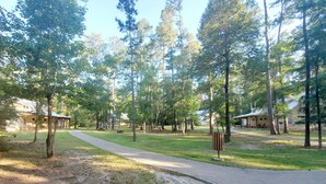 A park with a paved walkway, trees, and buildings with a rustic design. - YMCA Camp Cullen (Trinity)