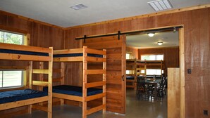 A bunk room with wooden walls and a dining area in the background. - YMCA Camp Cullen (Trinity)