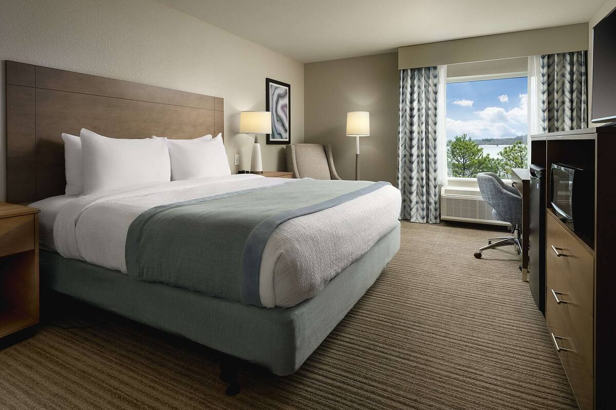 A hotel room with a large bed, two bedside tables, a desk with a chair, a television, and a view of the outdoors.