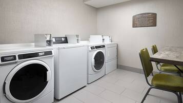 Une buanderie moderne équipée de deux machines à laver et deux sèche-linge Maytag, d’une table avec quatre chaises vertes et d’un panneau indiquant qu’elle est réservée à l’usage des clients.