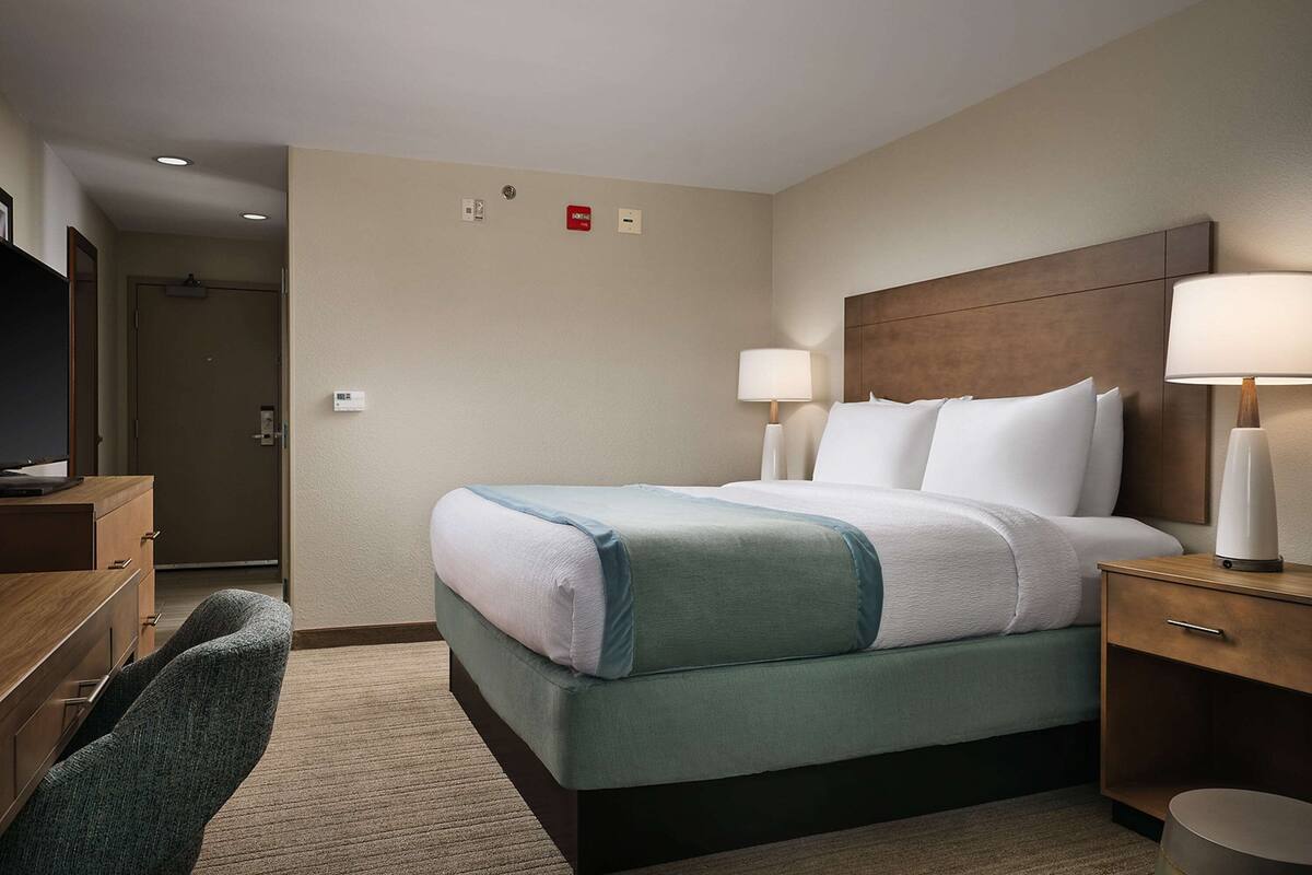 A hotel room with a bed, bedside lamps, a wooden nightstand, a desk, and a chair.