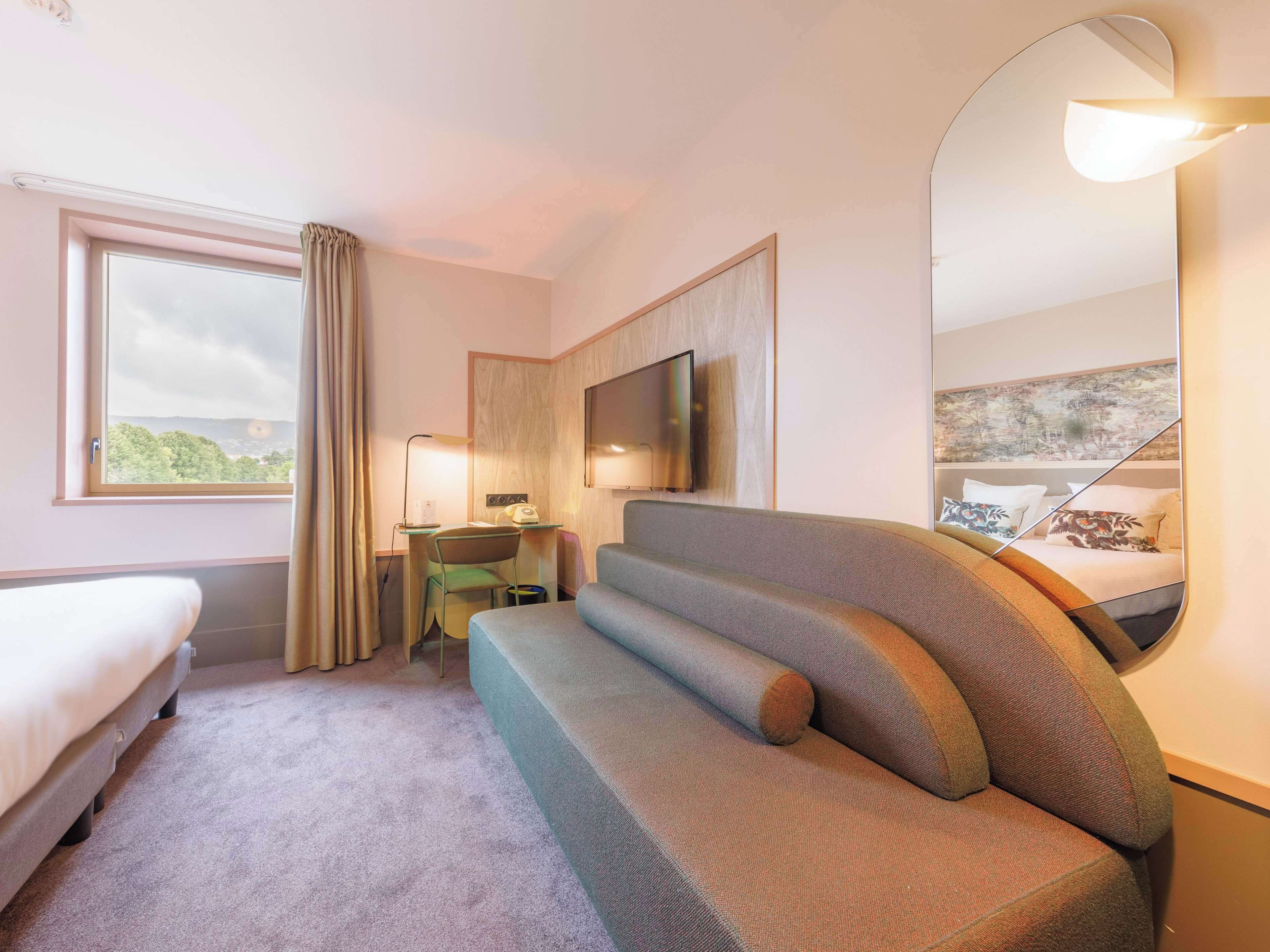 A hotel room with a bed, a sofa, a desk with a chair, a flat-screen TV, and a large window with curtains.