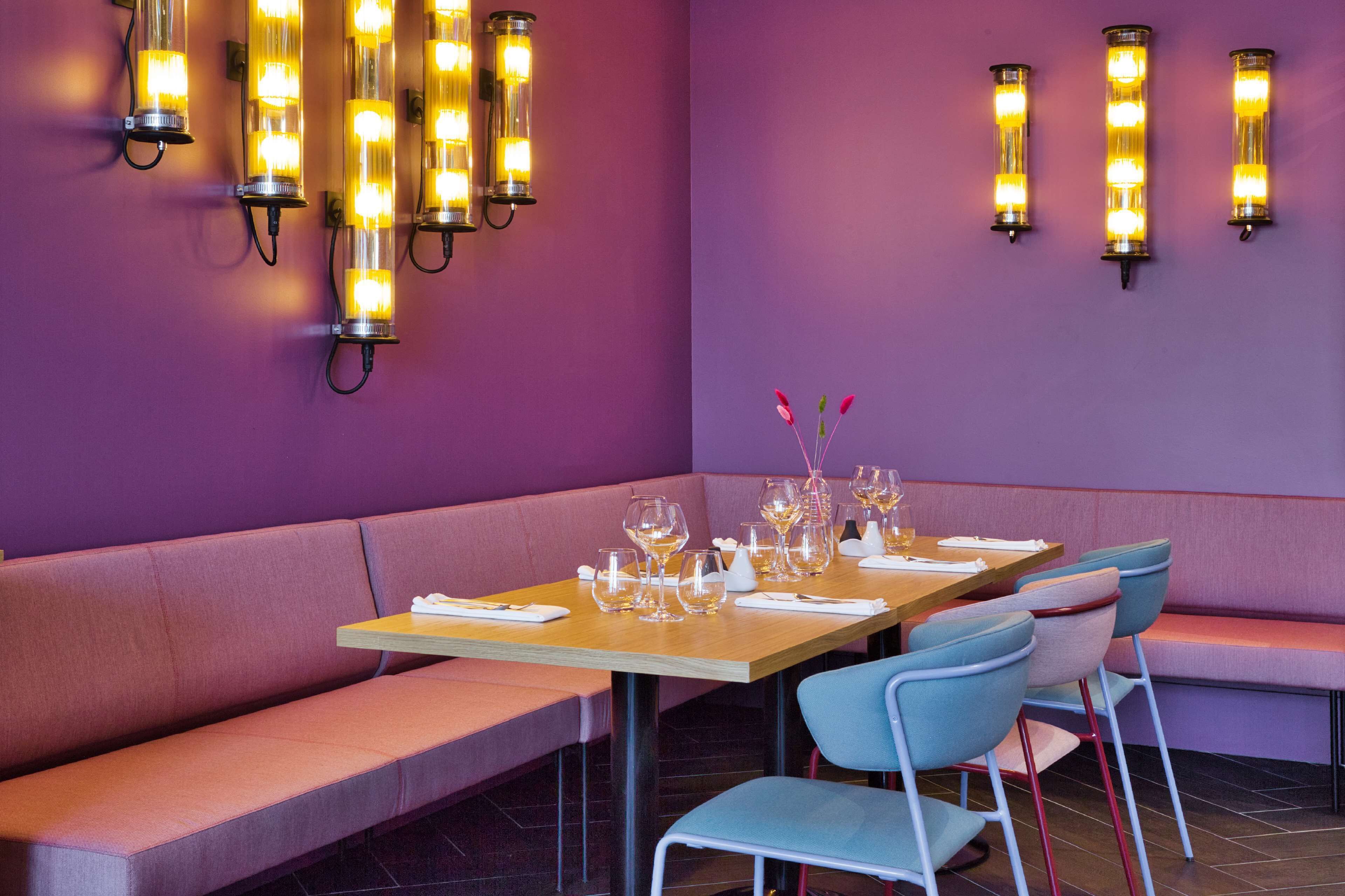 A restaurant interior with a purple wall, a wooden table set with wine glasses, and a bench.