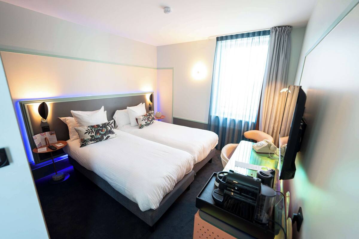 Ein Hotelzimmer mit zwei Betten, einem Schreibtisch mit Telefon und Kaffeemaschine sowie einem Fenster mit Vorhängen.