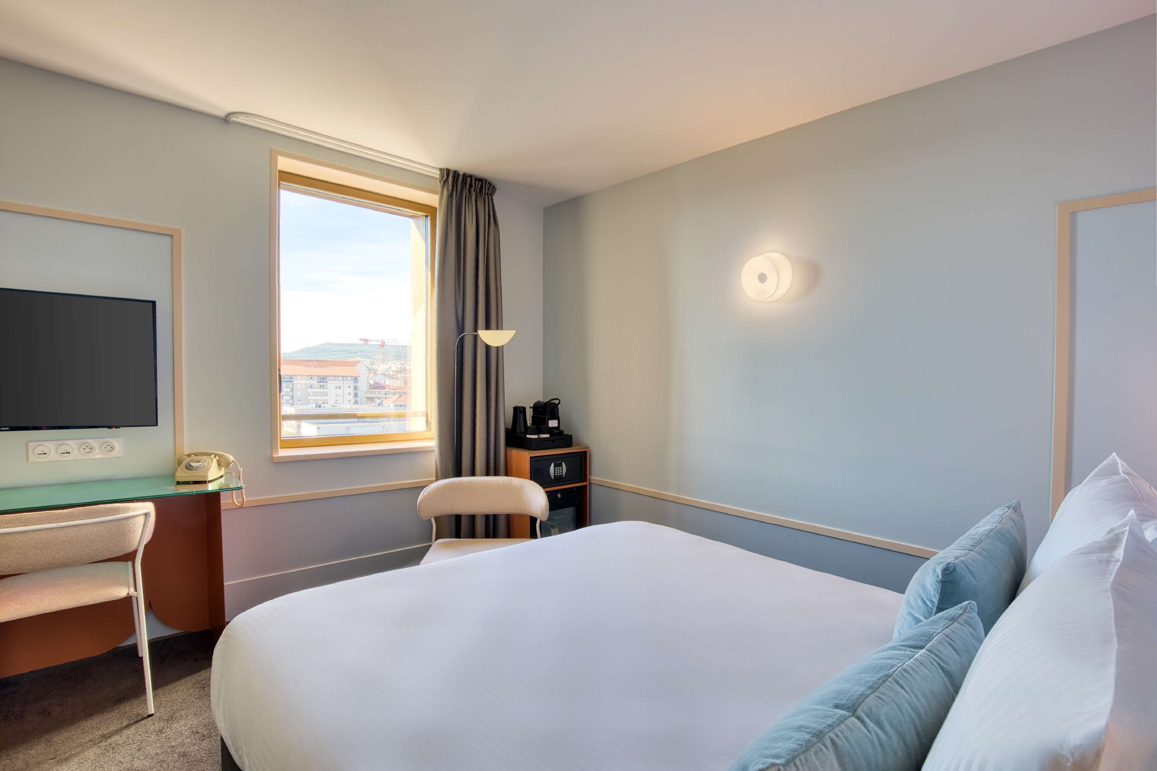 A hotel room with a bed, a desk with a chair, a television, a window with curtains, and a nightstand with a lamp.