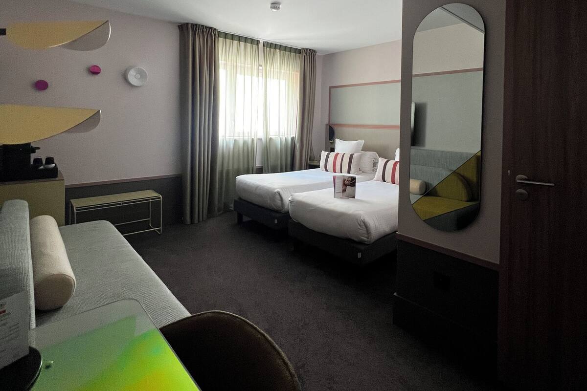 Ein Hotelzimmer mit zwei Betten, einem Schreibtisch, einem Stuhl, einem Spiegel und einem Fenster mit Vorhängen.