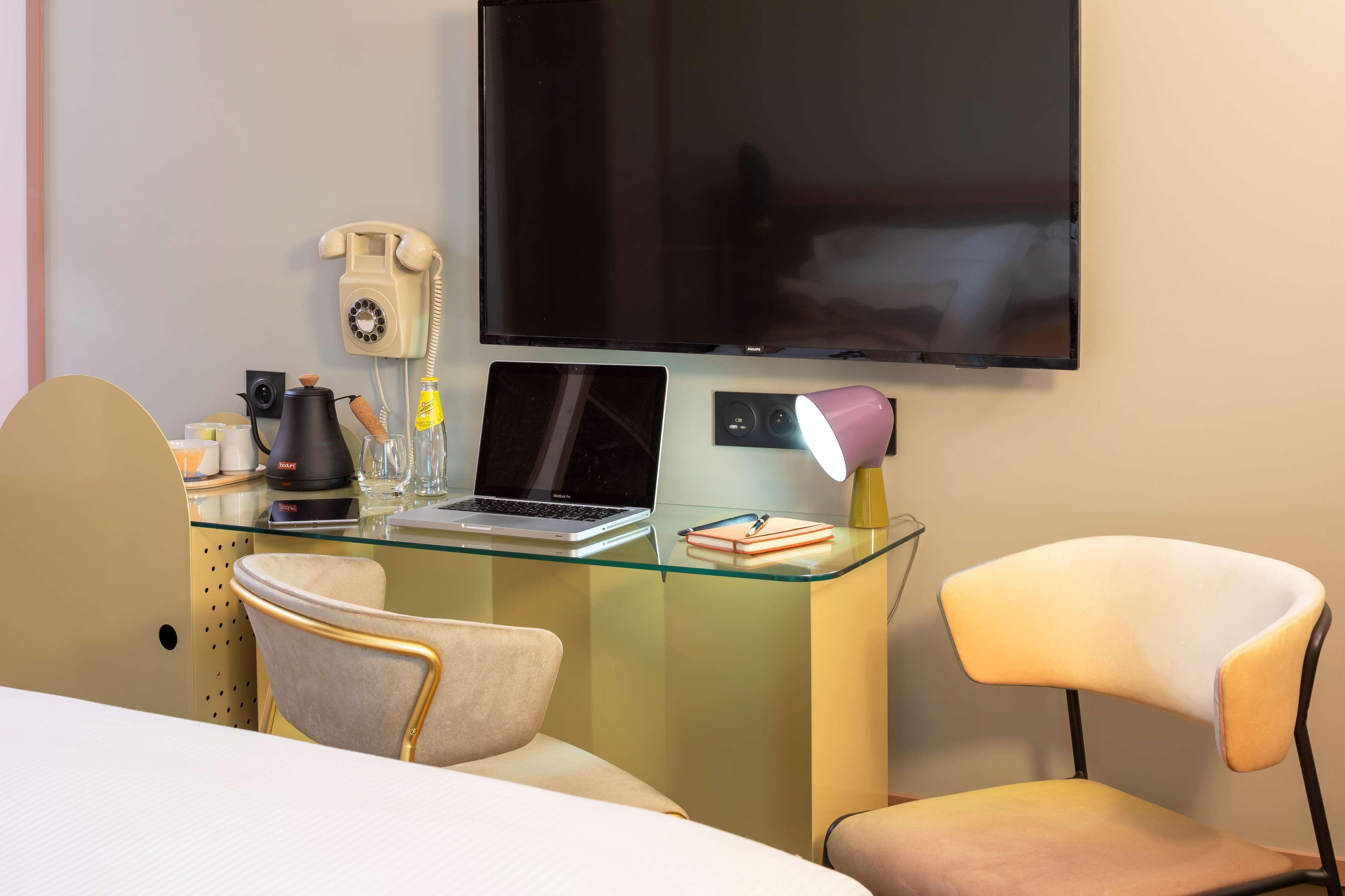 Een hotelkamer met een glazen bureau, een laptop, een telefoon, een televisie en twee stoelen.