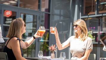 Two people toasting with drinks at an outdoor table with a glass vase of flowers.