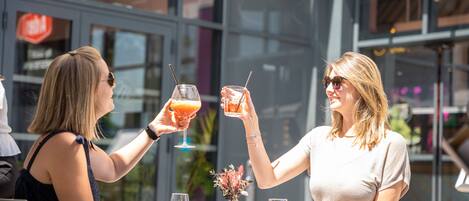 Two people toasting with drinks at an outdoor table with a glass vase of flowers.