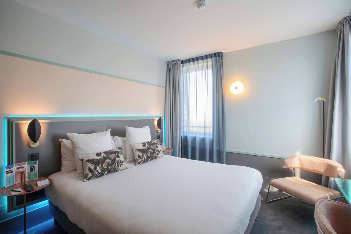 Ein Hotelzimmer mit einem großen Bett, einem Nachttisch mit Lampe, einem Sessel und einem Fenster mit Vorhängen.