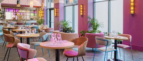A modern restaurant with round tables and chairs, large windows, and decorative wall lights.