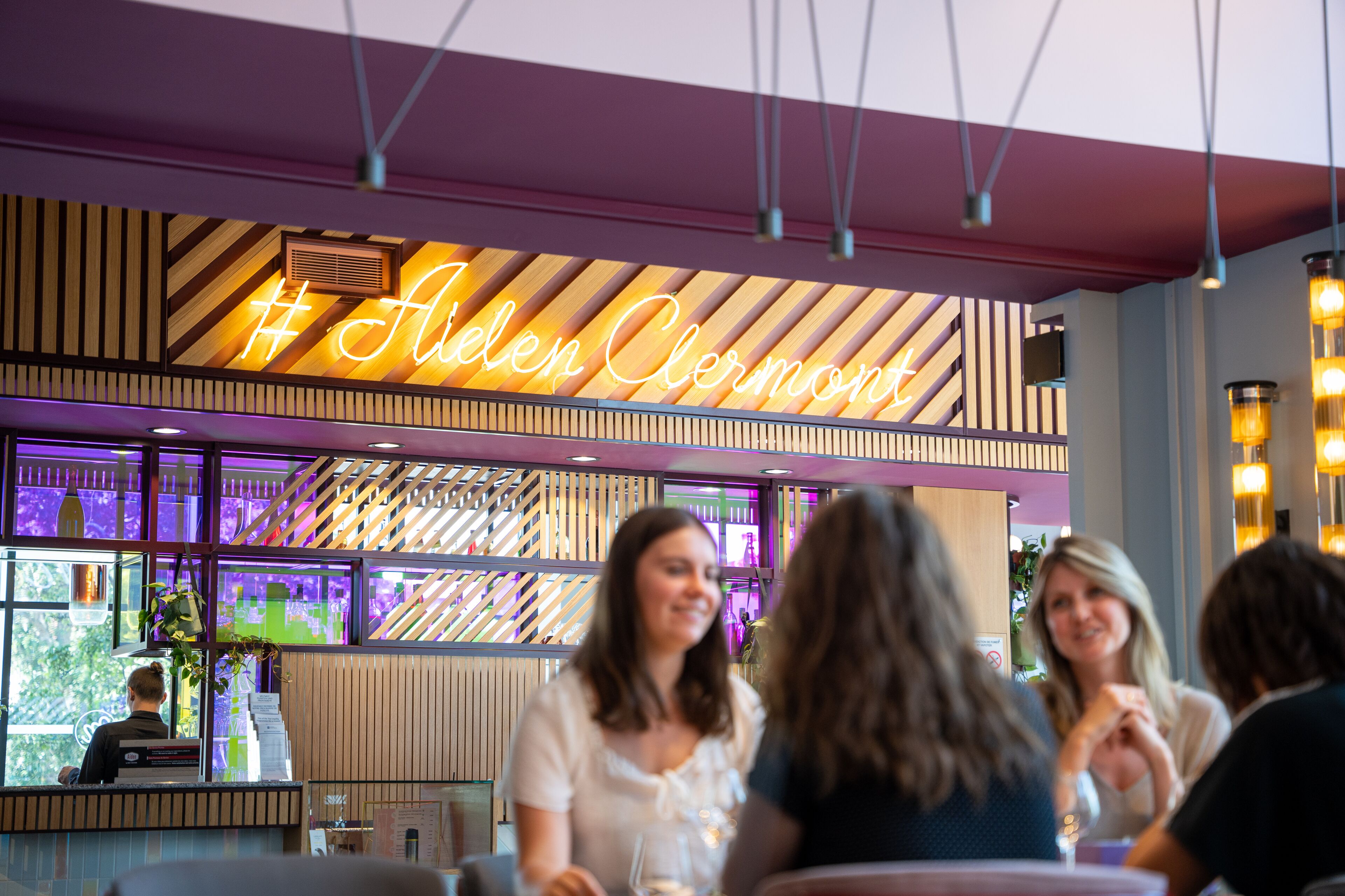 Interior de un restaurante con un letrero de neón que dice '#Alden Clermont' y un diseño de techo moderno a rayas.