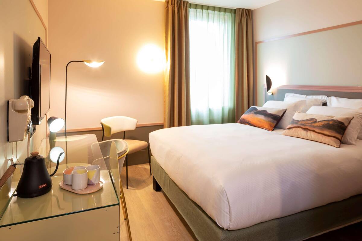 Ein Hotelzimmer mit Bett, Nachttisch, Lampe und einem Fenster mit Vorhängen.
