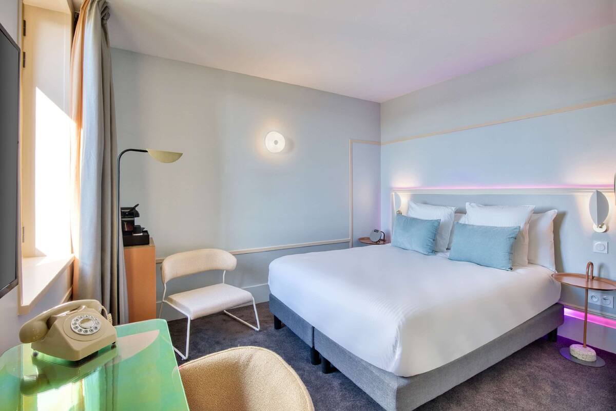 Ein modernes Hotelzimmer mit einem großen Bett, einem Schreibtisch mit Telefon, einem Sessel und einem Nachttisch mit Lampe.