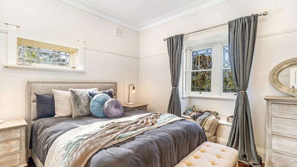 A bedroom with a large bed, a nightstand, a dresser, a window with curtains, and a stained glass window above the bed. - Berry Bliss, Berry, South Coast (Berry)