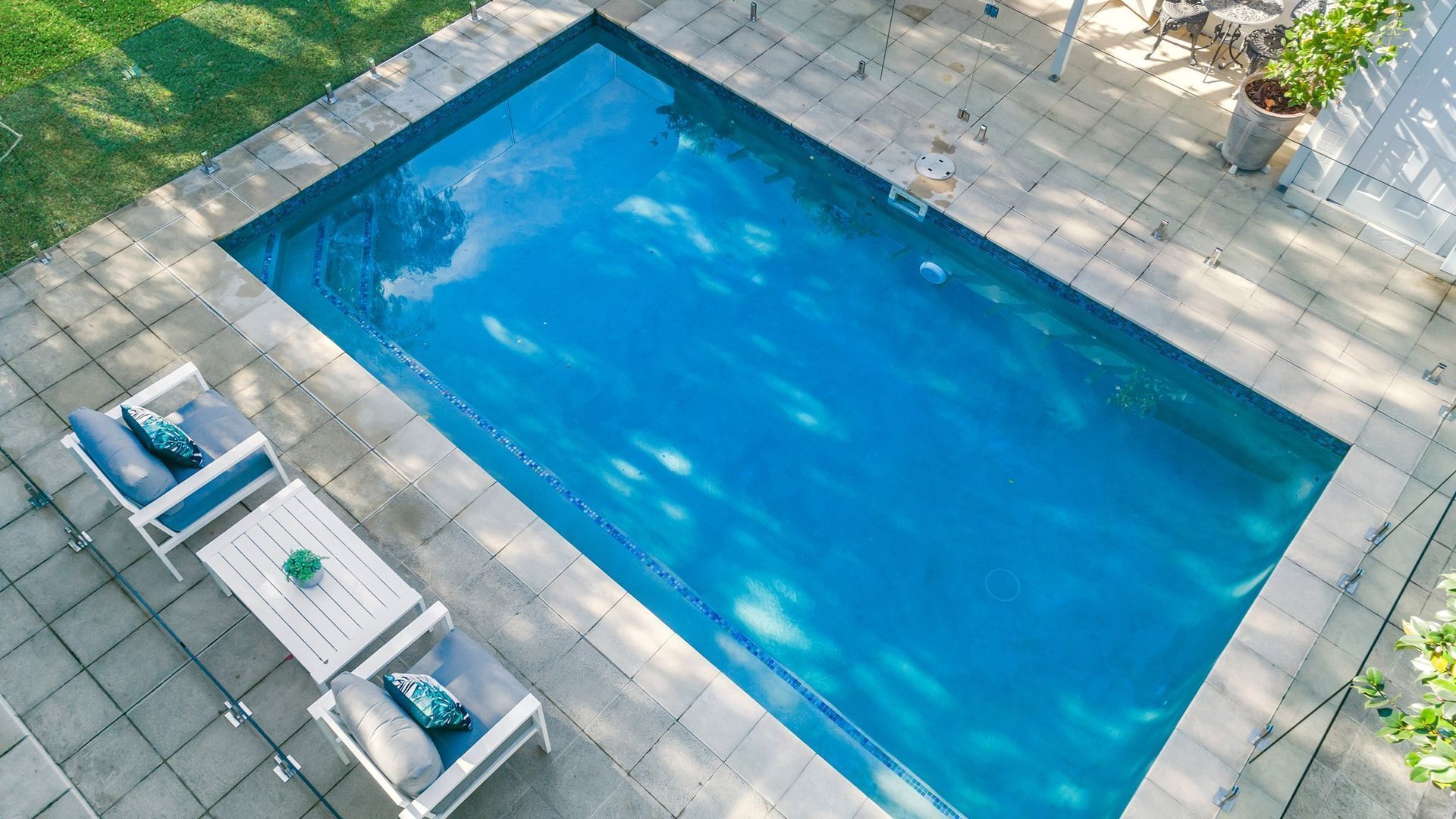 A rectangular swimming pool with a tiled deck, surrounded by a grassy area and a few potted plants.