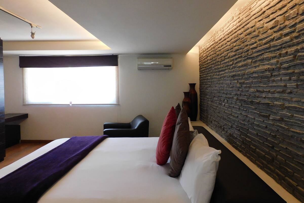 A modern hotel room with a large bed, a window, a brick accent wall, and a black chair.