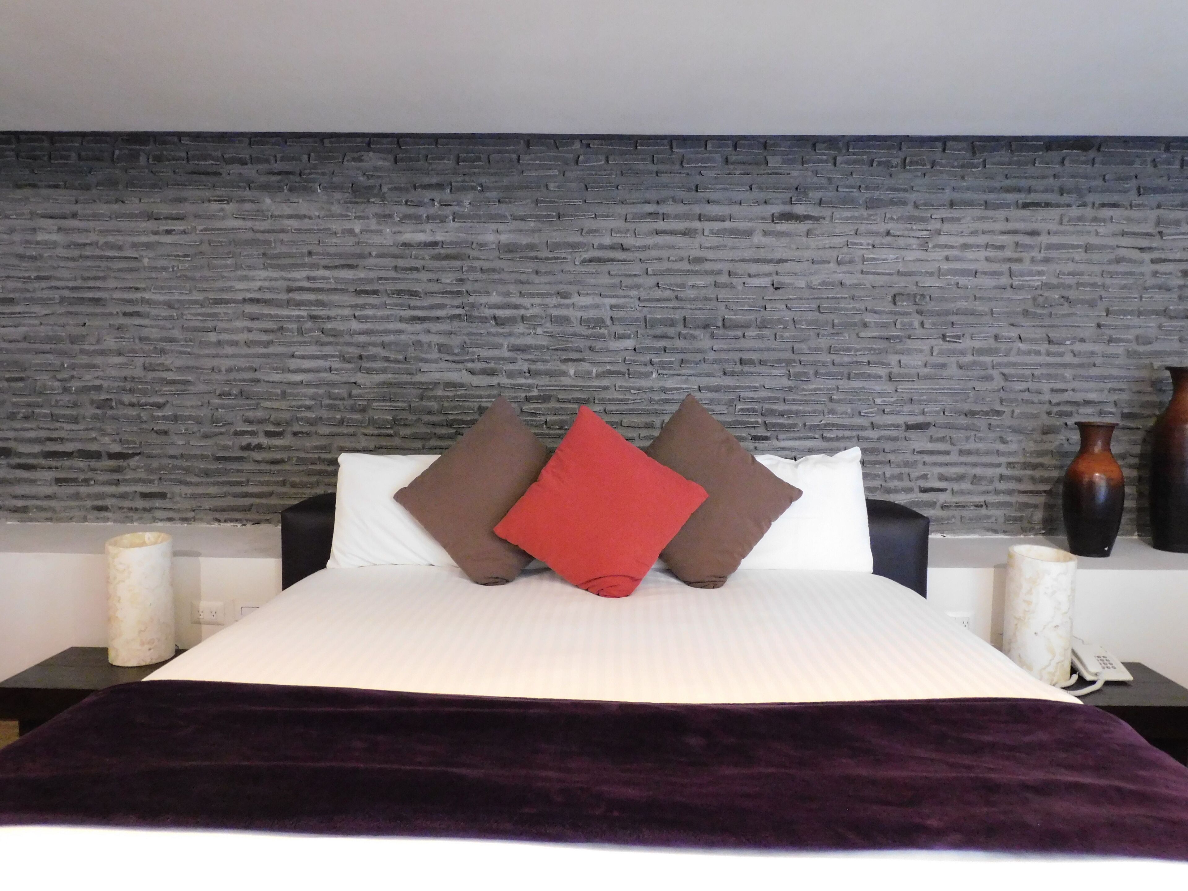 A bed with white bedding, a dark purple throw blanket, and three pillows in brown and red.