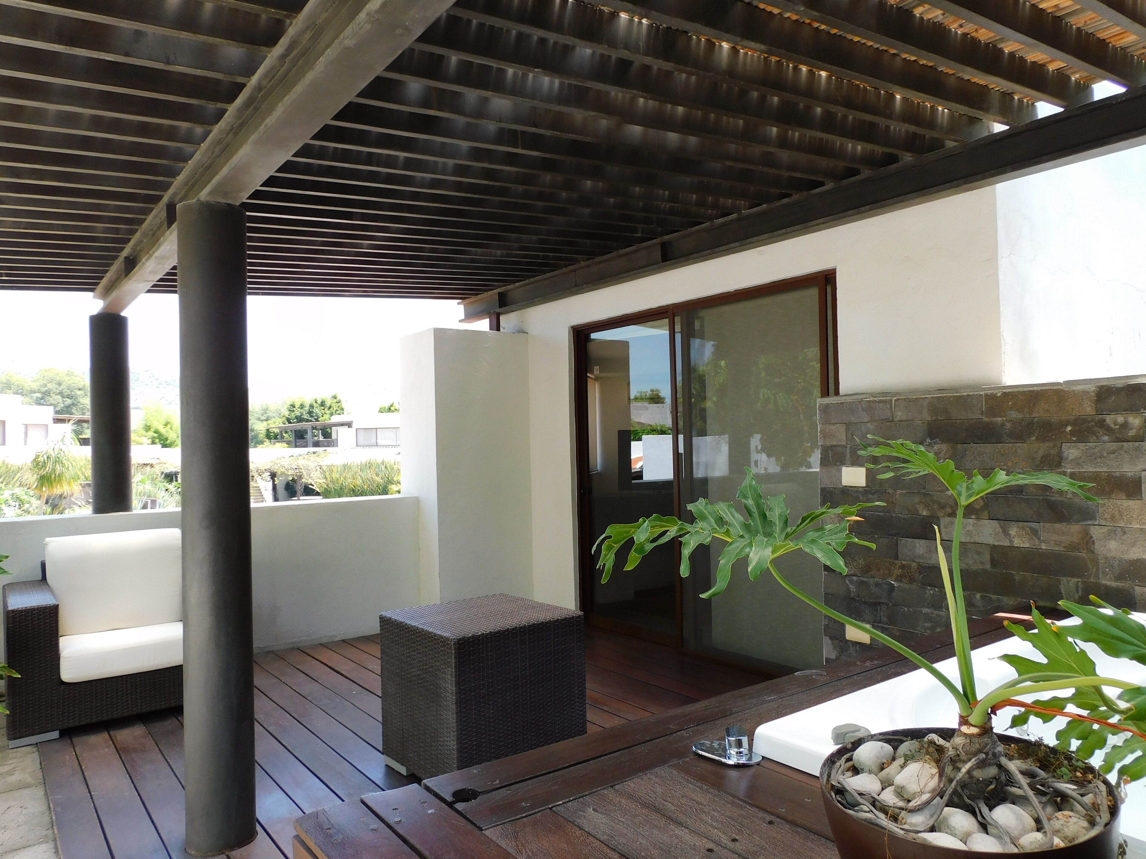 A covered outdoor area with a wooden deck, a white sofa, a wicker chair, and a potted plant.