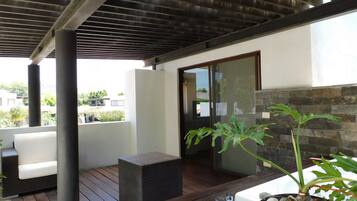 A covered outdoor area with a wooden deck, a white sofa, a wicker chair, and a potted plant.