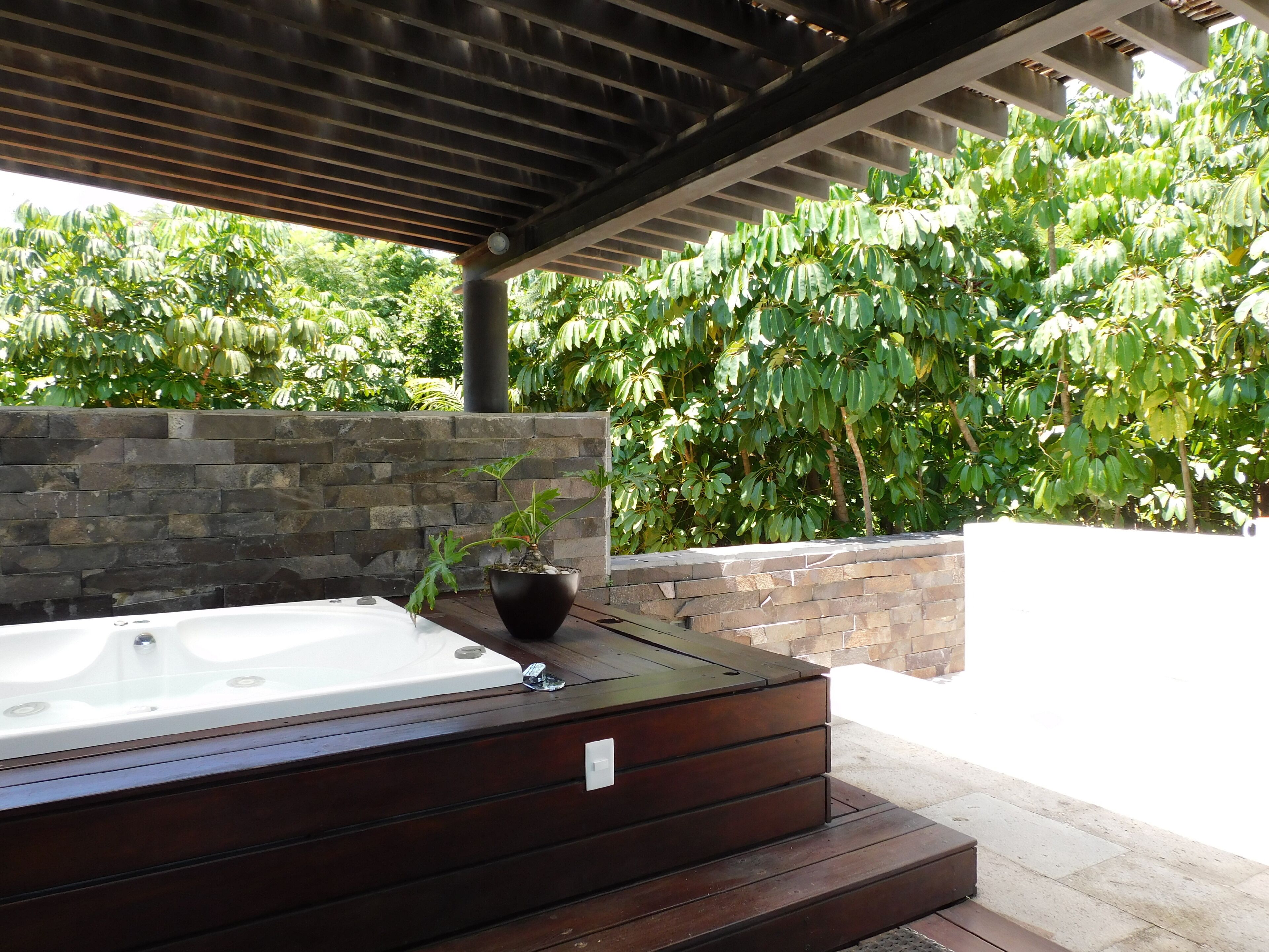 A hot tub under a wooden pergola with a stone wall and lush greenery in the background.