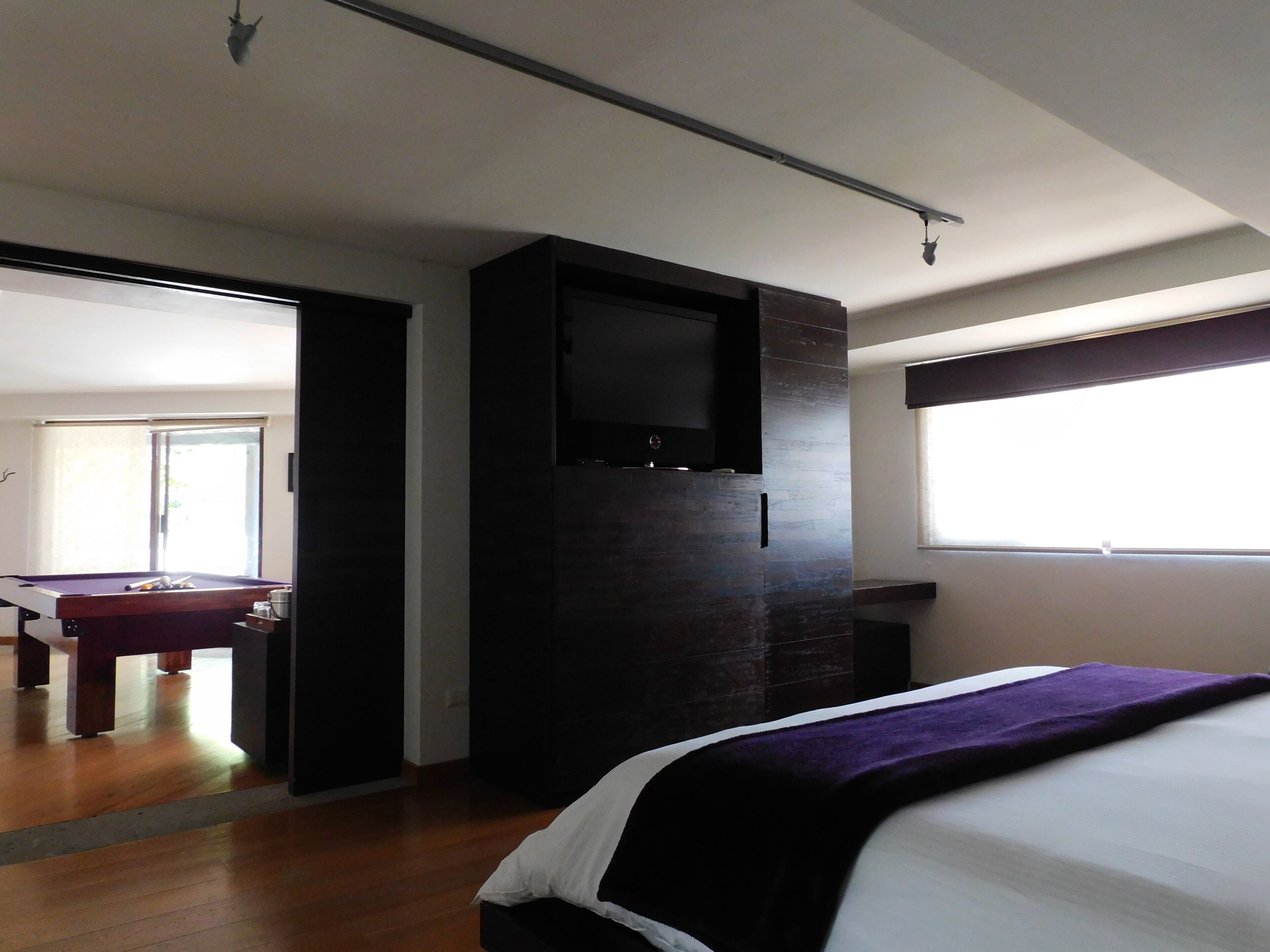 A hotel room with a bed, a flat-screen TV mounted on a dark wall, a pool table, and a window with blinds.