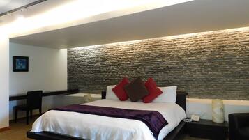 Un dormitorio moderno con pared de piedra, cama con ropa de cama blanca y varios almohadones, un escritorio y una silla.