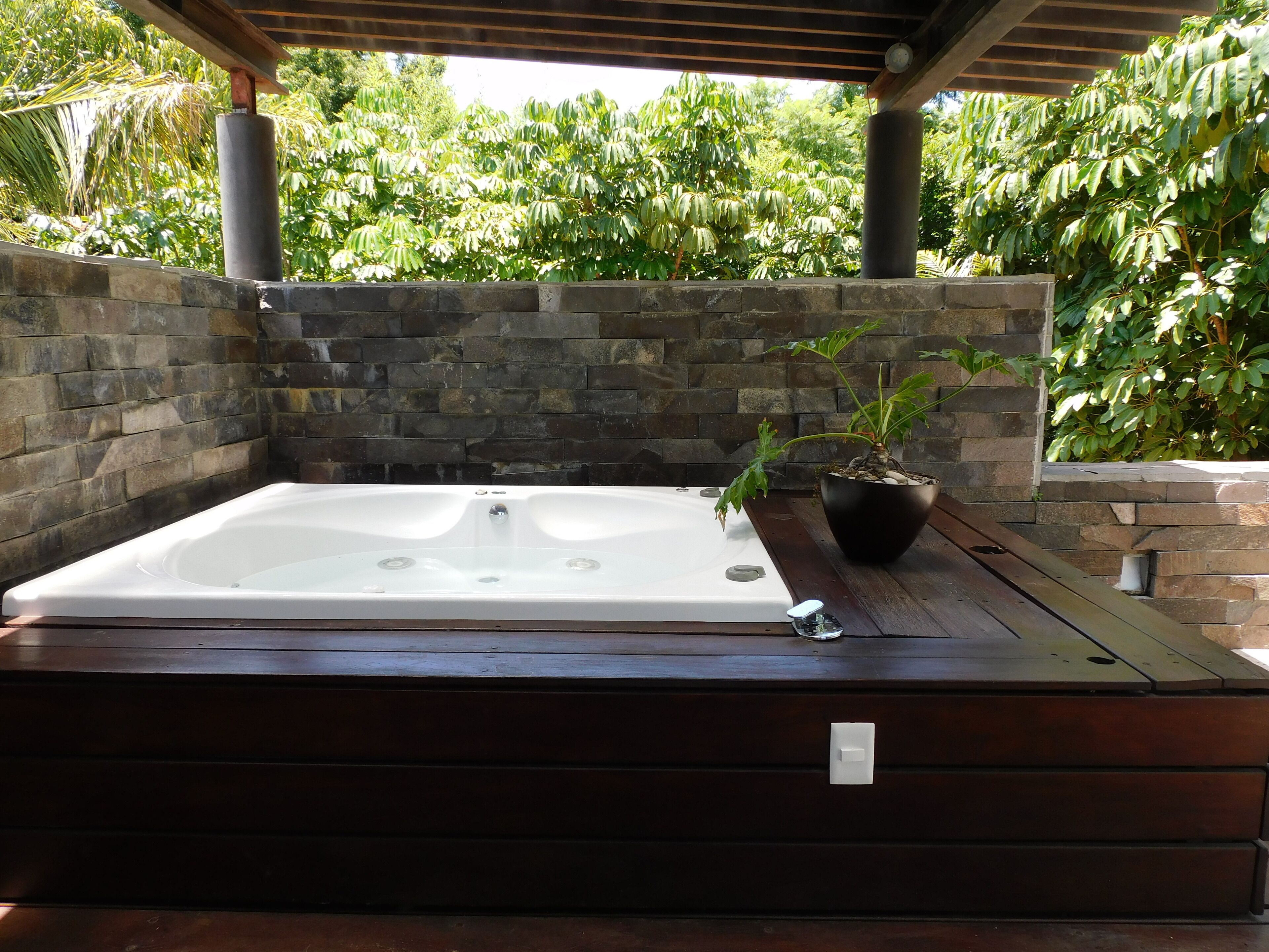 Un jacuzzi bajo una pérgola de madera, con paredes de piedra y abundante vegetación al fondo.