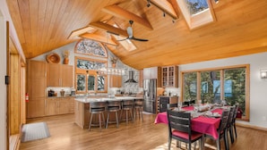 A spacious kitchen with wooden cabinets, a central island with stools, a dining area with a table set for six, and large windows offering views of the outdoors.