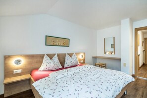 Ein Schlafzimmer mit Holz-Kopfteil, einem Bett mit floralem Bettbezug, Nachttischlampen, einem kleinen Nachttisch, einem gerahmten Bild an der Wand und einem Spiegel über dem Bett.