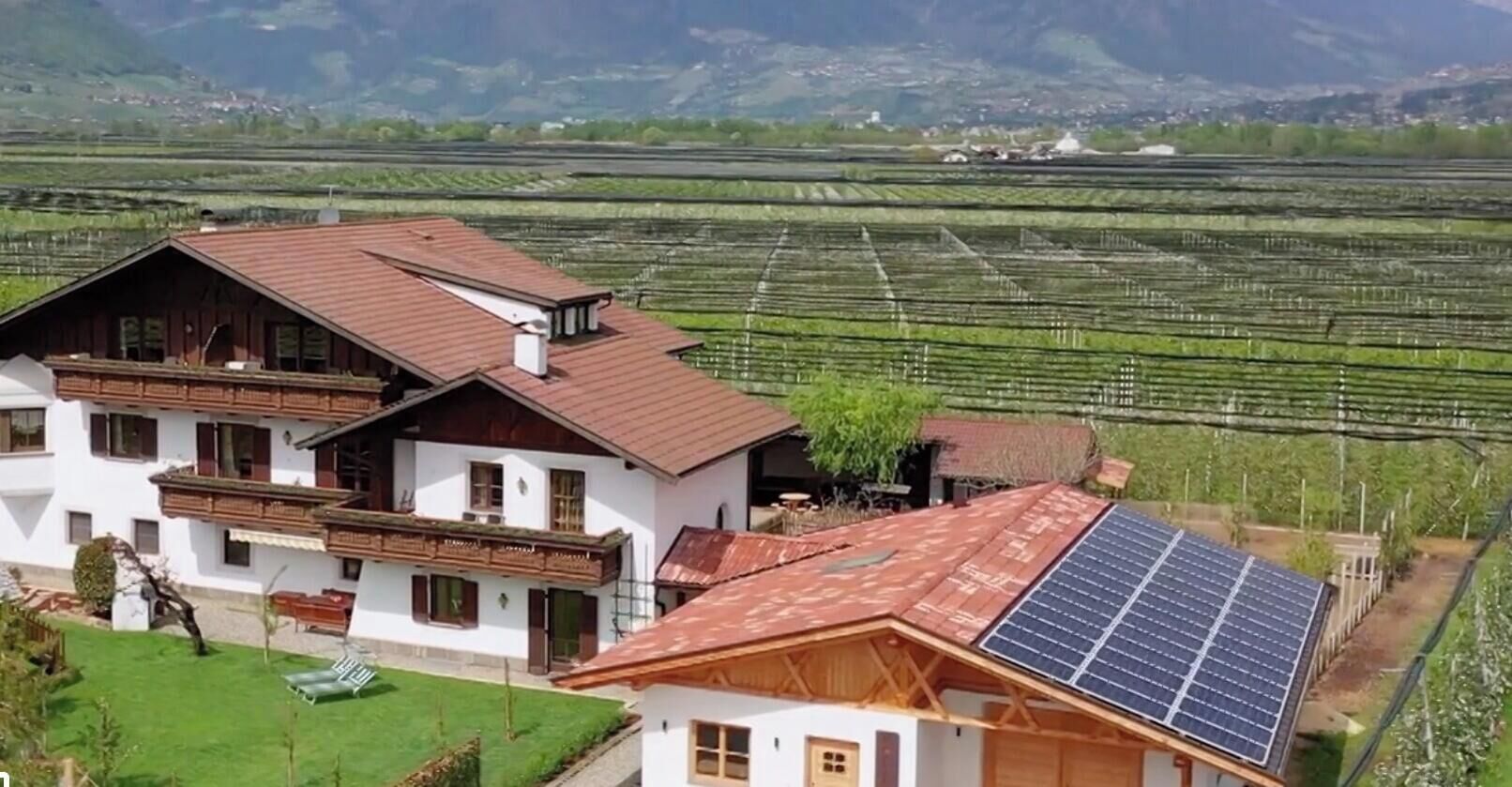 Una casa a due piani con tetto spiovente, un pannello solare sul lato destro e un vigneto sullo sfondo.