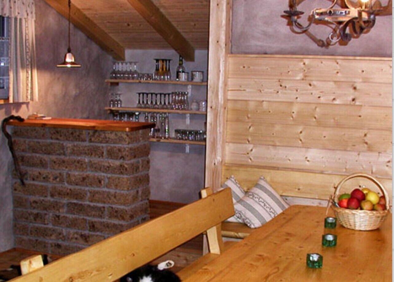 Ein rustikaler Holzinnenraum mit Kamin aus Ziegelsteinen, hölzernen Bänken, einem Tisch mit Obstkorb und Regalen mit Glaswaren.