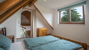 Camera da letto con soffitto spiovente, struttura del letto in legno, finestra vista alberi e piccolo mobiletto in legno.