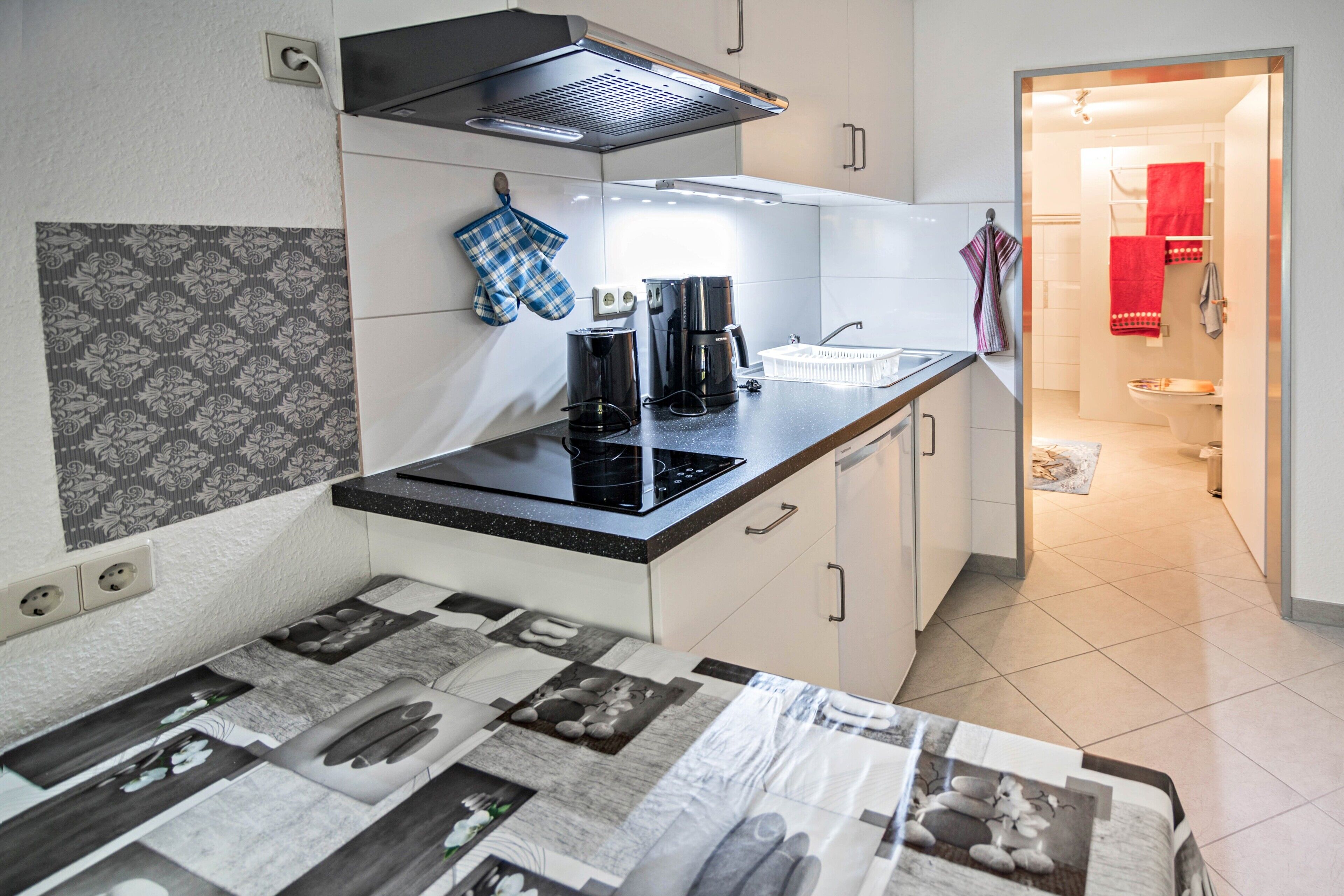 Eine kleine Küche mit Herd, Kaffeemaschine und einer Arbeitsplatte. Rückwand mit Muster und geflieste Böden.