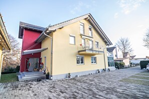A two-story house with a red and yellow facade, a balcony, and a paved driveway.