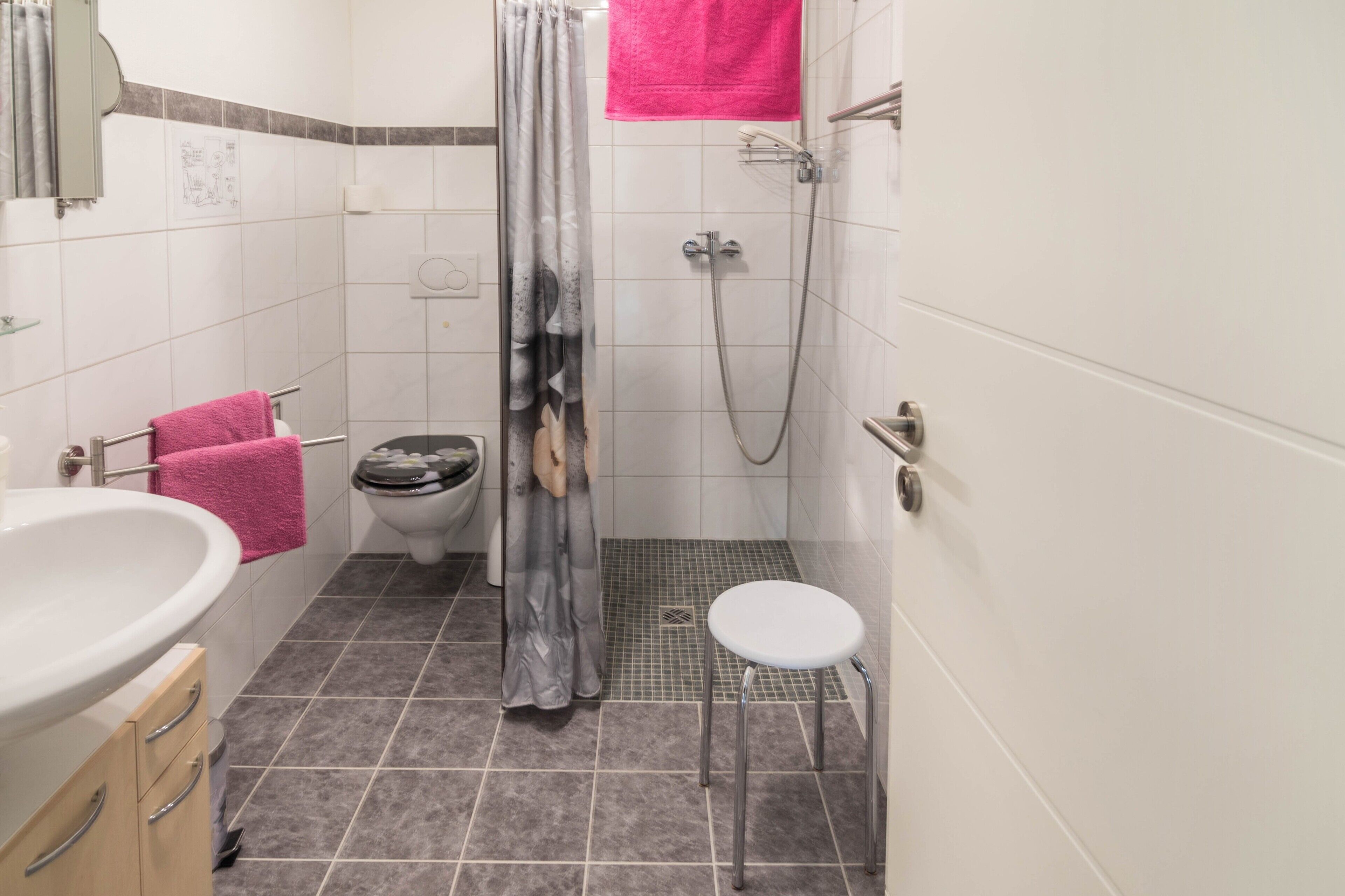 A bathroom with a shower, toilet, sink, and a small stool.