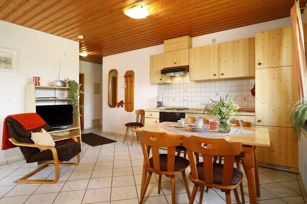 A modern kitchen with wooden cabinets, a dining table, and a TV in a living area with a rocking chair.