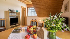 A dining area with a wooden table, a fruit basket, a vase with flowers, and a television on a shelf. - Holiday Apartment 1 on “Wagenberg” Farm with Mountain View, Wi-Fi & Garden (Deggenhausertal)