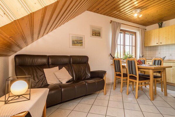 A cozy living room with a leather sofa, a dining table with chairs, wooden cabinets, and a window with curtains.