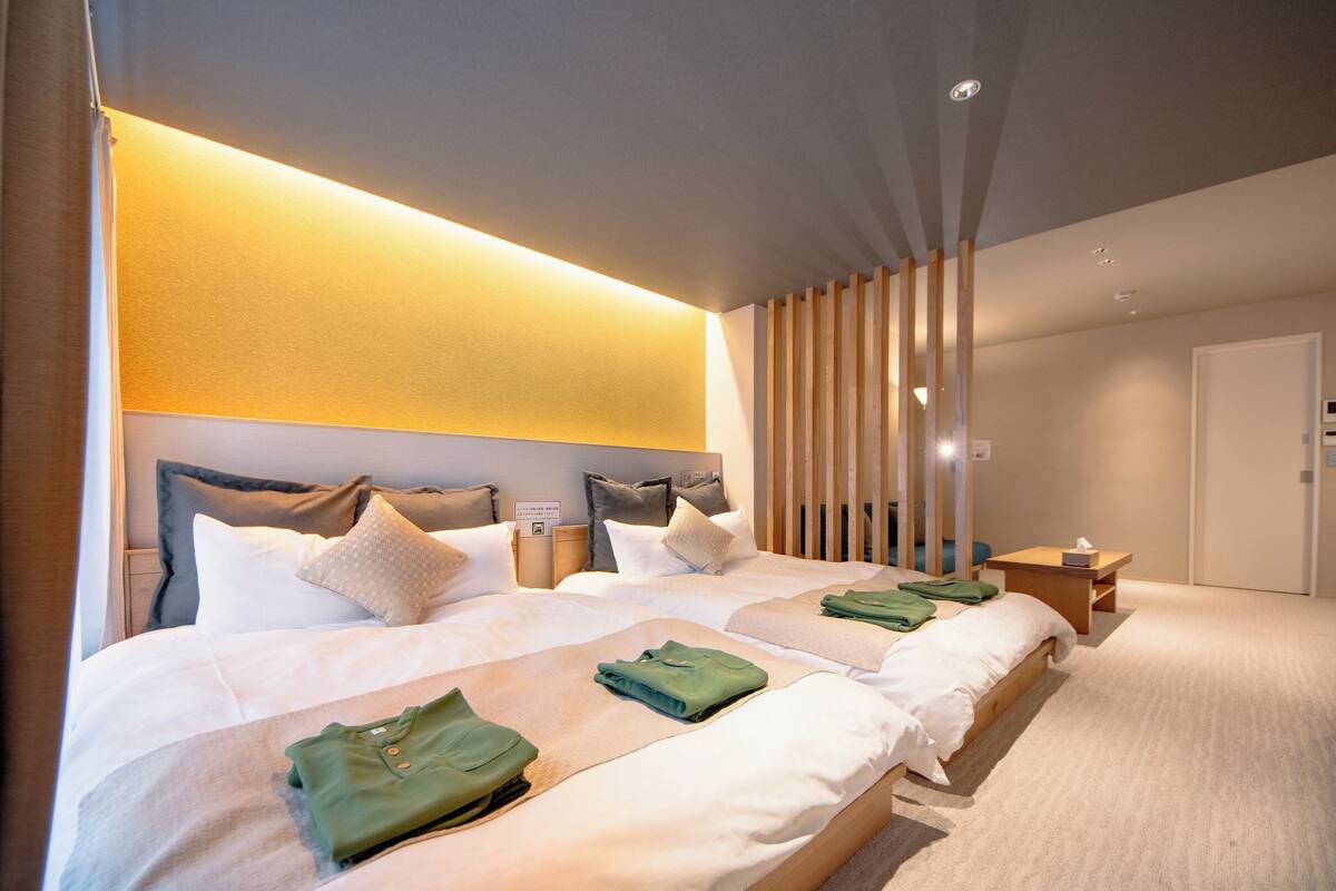 A modern hotel room with two beds, a wooden headboard, a bedside table, and a wall-mounted light fixture.