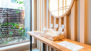 A reception desk with a wooden finish, a round window, and a view of a brick wall and greenery outside. - Hotel Rokune (Nara)
