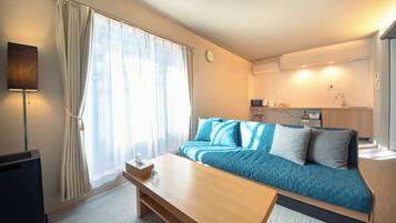 Una habitación de hotel moderna con sofá cama, una mesa de madera, un televisor, una ventana con cortinas y una zona de kitchenette.
