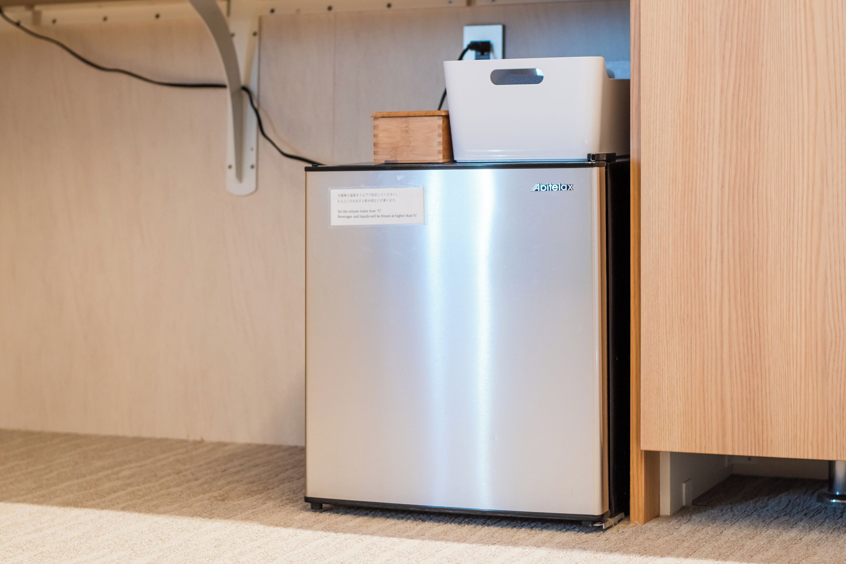 A compact, stainless steel refrigerator with a note on its door, placed in a room with a wooden cabinet and a carpeted floor.